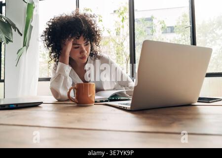 Hispanic woman sitting at table touching forehead during work at home Stock Photo
