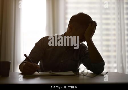 Depressed Asian male writing on book leaning head on hand while sitting at table in office Stock Photo