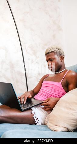 Black non-binary person using laptop sitting on sofa in living room at home Stock Photo