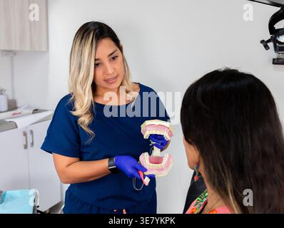 Mixed race female dentist wearing latex gloves demonstrating dental hygiene using toothbrush and model of teeth to Hispanic female patient sitting ... Stock Photo