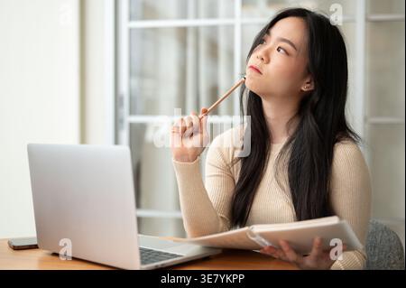 Serious Asian woman  looking away holding pen and notebook sitting at table with laptop at home Stock Photo