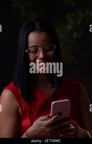 Smiling Hispanic woman using smartphone standing against dark background Stock Photo