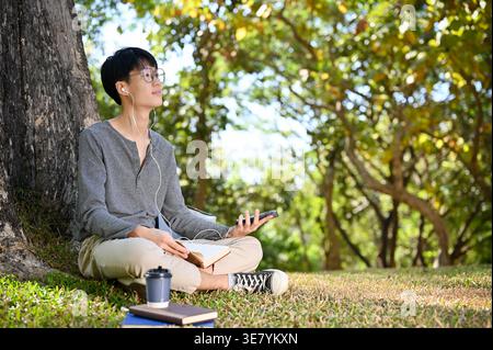 Asian male in earphones holding smartphone looking away while sitting on grass in park on sunny day Stock Photo