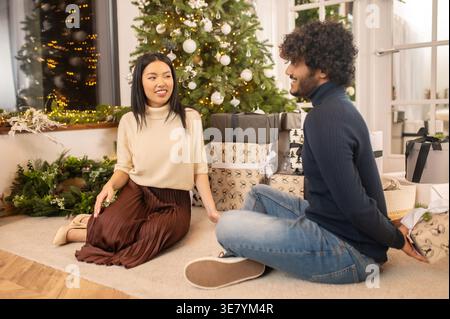 Asian man hiding gift behind sitting with girlfriend on floor during Christmas Stock Photo