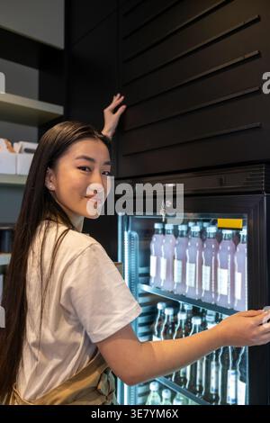 Smiling Asian female cafe worker standing on the stepladder and touching the wall over the refrigerated display cabinet with soft drinks in cafe Stock Photo