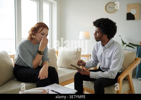 Black male psychologist with afro taking notes while talking to Asian female patient wiping tears sitting together on wooden couch during consultat... Stock Photo
