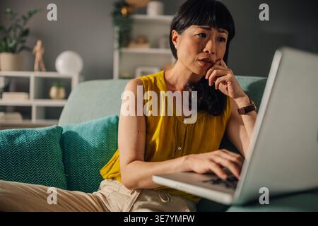 Japanese woman with hand on face using laptop while sitting with crossed legs on sofa in living room at home Stock Photo