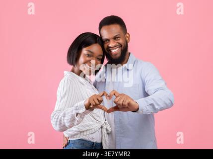 Smiling Black couple making heart with hands while standing against pink background in studio Stock Photo