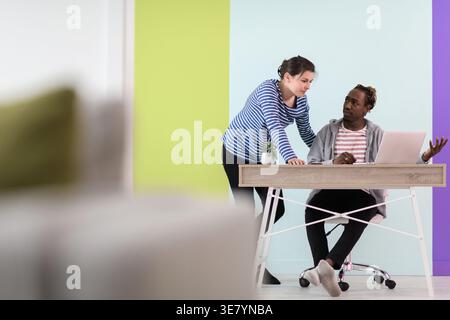 Black businessman with braids sitting at table working on laptop and having discussion with caucasian female colleague standing beside him in the o... Stock Photo