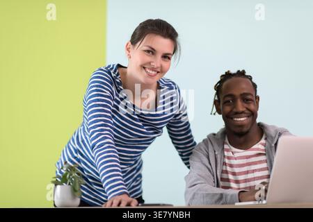 Smiling Black businessman with braids sitting at table with laptop near caucasian female colleague standing beside him against colorful wall in the... Stock Photo