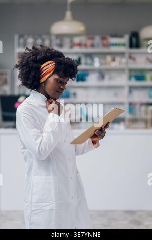Black female pharmacist with afro touching neck and holding clipboard standing inside pharmacy Stock Photo