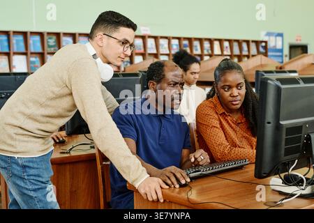 Middle Eastern man with headphones around neck helping middle-aged Black man and Black woman in using computer in university library Stock Photo