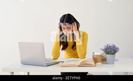 Tired Asian woman wearing yellow shirt with eyeglasses on forehead touching head while sitting at table in office Stock Photo