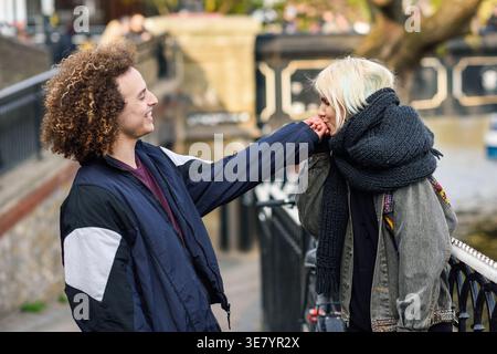 Smiling Mixed race man with curly hair looking at caucasian girlfriend kissing his hand while standing on city street during daytime Stock Photo