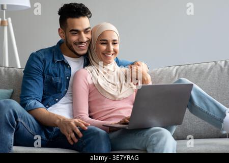 Smiling Middle Eastern couple using laptop while sitting close together on sofa at home Stock Photo