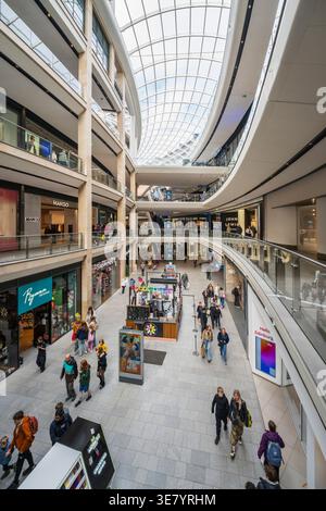People walk through a bright, multi-level shopping mall with a distinctive curved glass ceiling and various retail stores Stock Photo