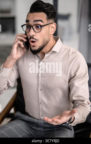 Surprised Arab man wearing eyeglasses having phone conversation sitting at table in office Stock Photo
