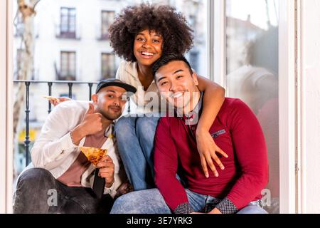 Smiling Black woman with afro holding pizza and hugging male friends while sitting together on balcony doorway at home Stock Photo
