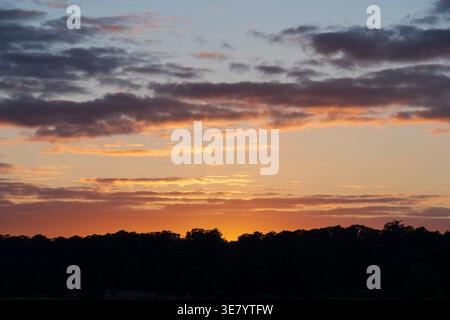 The sun dips below the horizon, painting the sky with fiery hues and casting a deep shadow over the treeline Stock Photo