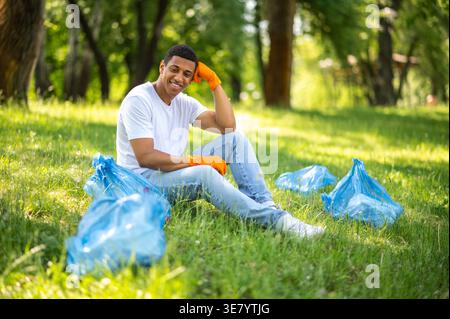 Smiling Black man wearing orange gloves touching head sitting on grass near plastic bag in park on sunny day Stock Photo