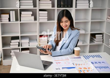 Smiling Asian business woman holding calculator working on business financial data while sitting at table in office Stock Photo