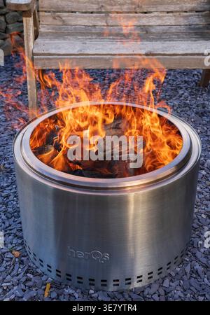 A stainless steel fire pit burns brightly with logs inside, set on a gravel surface with a wooden bench nearby Stock Photo