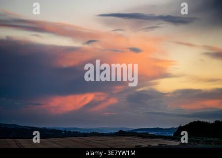 A breathtaking sunset unfolds with fiery clouds casting a warm glow over rolling hills and harvested fields below Stock Photo