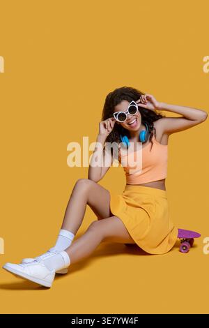 Smiling African American female touching sunglasses sitting on skateboard against yellow background Stock Photo