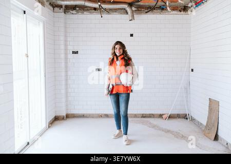 Smiling Hispanic female architect wearing safety vest holding clipboard and hard hat standing in construction site during daytime Stock Photo