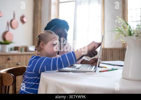 Smiling Black female tutor with afro sitting at table with caucasian girl with down syndrome and having a lesson using laptop at home Stock Photo