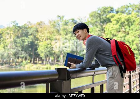 Asian male student holding books looking away while standing with backpack and leaning on metal railing in park during daytime Stock Photo