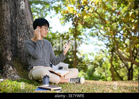 Asian male touching earphones using smartphone while sitting on grass in park on sunny day Stock Photo