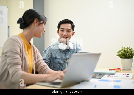 Smiling Asian male UX graphic designer with headphones around neck looking and discussing work with his female colleague while sitting at table in ... Stock Photo