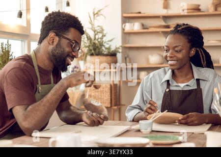 Black male artist wearing apron and eyeglasses holding brush and palette explaining pottery details with female student sitting at table in workshop Stock Photo