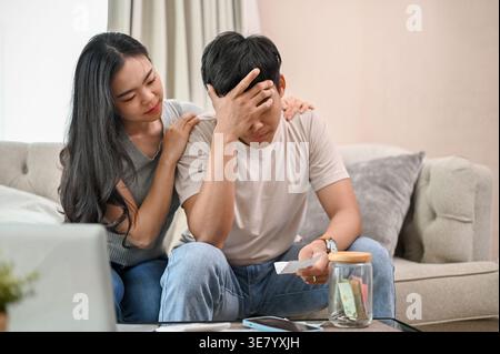 Stressed Asian couple looking at document sitting on sofa near table with jar of money in living room at home Stock Photo