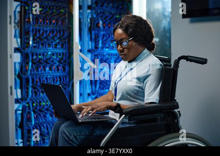 Mixed-race woman with eyeglasses using laptop while sitting on wheelchair in server room Stock Photo