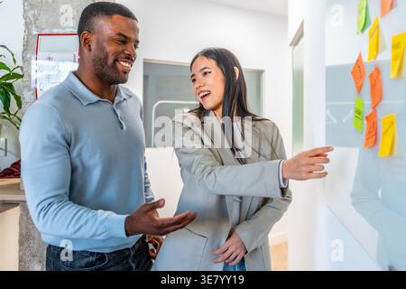 Black male and Asian female colleagues discussing business strategy on whiteboard using sticky notes while standing together in office Stock Photo