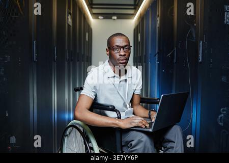 Disabled Black male IT engineer sitting on wheelchair with laptop in server room in data center Stock Photo