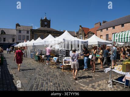 People are browsing artwork displayed in tents at a sunny outdoor market in a cobblestone town square with historic buildings in the background Stock Photo