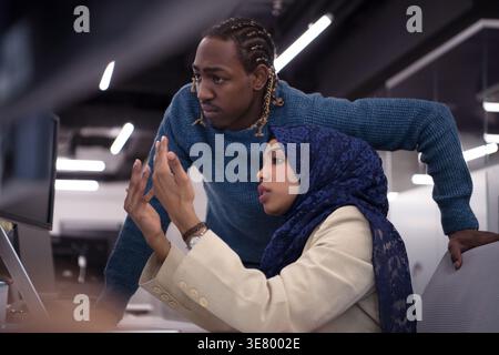 Black male employee standing beside female colleague leaning arms with open palms on table discussing and using desktop sitting at table in office Stock Photo