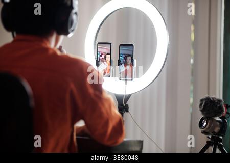 Asian male online seller wearing headset showing shoe sitting at table broadcasting live on two smartphones in front of ring light and recording on... Stock Photo