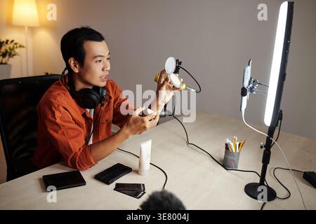 Asian male online seller showing foundation to camera sitting at table with makeup and skincare products streaming live from home studio Stock Photo