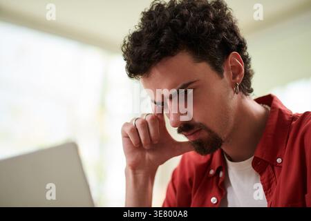 Mixed-race male office worker touching face looking at laptop sitting at table in office Stock Photo