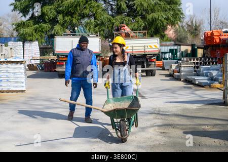 Happy Hispanic female worker wearing denim overalls, yellow hardhat, and protective gloves pushing wheelbarrow near Black foreman walking at constr... Stock Photo