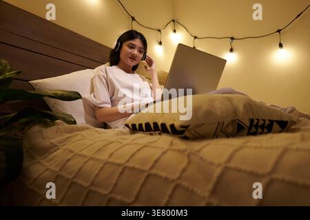 Smiling Asian woman wearing headphones watching webinar on laptop sitting on bed at home Stock Photo
