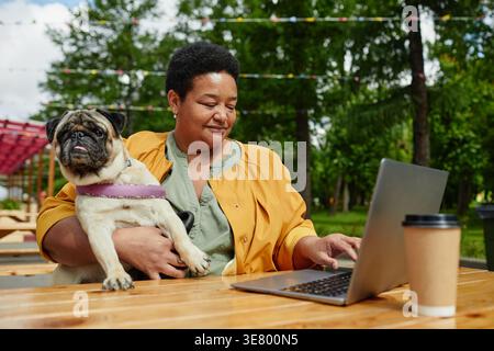 Black middle-aged woman working on laptop while holding her dog sitting at wooden table in outdoor cafe Stock Photo