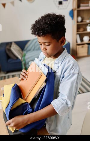 Black boy putting books on school bag standing at table at home Stock Photo