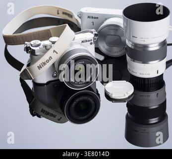 A white Nikon 1 mirrorless camera with a 1 NIKKOR lens, lens hood, and lens cap are displayed on a glossy black surface Stock Photo