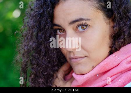 Serious Arab woman with curly hair wearing pink scarf standing in garden during daytime Stock Photo