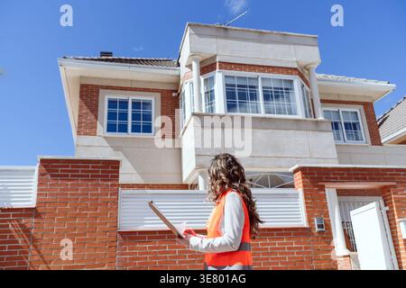 Hispanic female architect wearing vest holding clipboard standing on street checking house during daytime Stock Photo